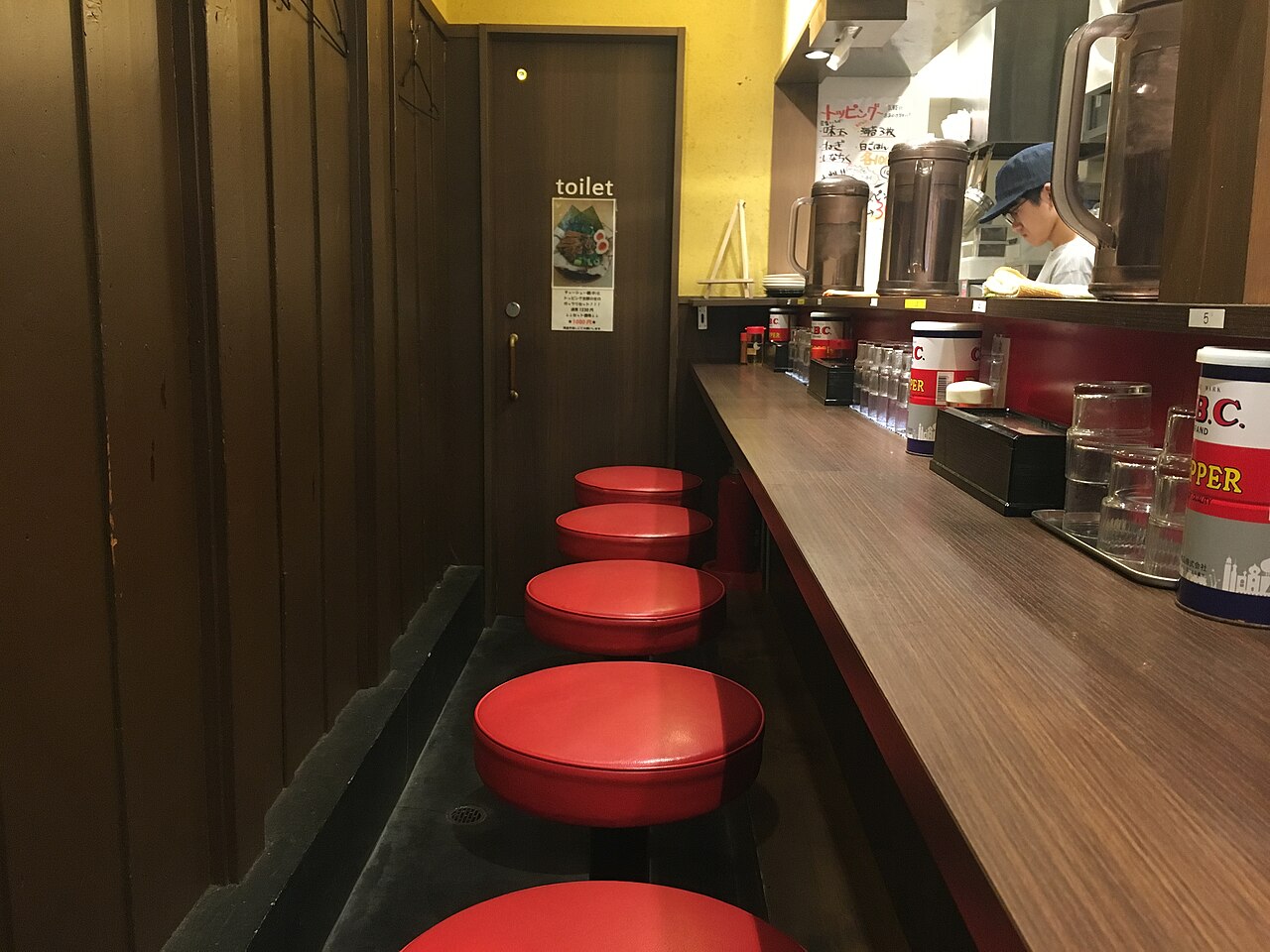 日本一間狹長拉麵店的吧台座位，一整排紅色圓凳沿著木質吧台排開，吧台上放著胡椒罐與水壺，內場戴著帽子的店員正在準備餐點